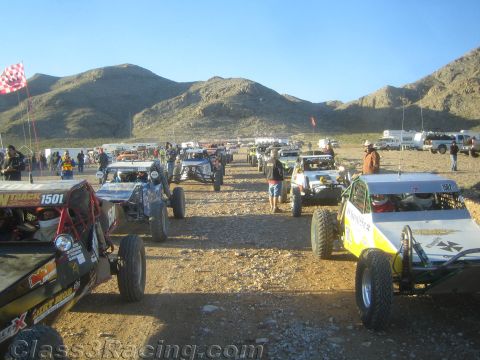 1990 Ford Bronco - long lineup of off-road buggies and trucks, including number 1501, fills a gravel staging area below desert mountains.