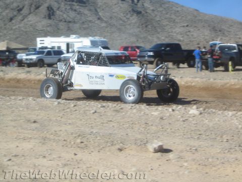 1990 Ford Bronco - white tube-frame off-road buggy lettered Tru Built races along a dirt track with vehicles and spectators in the background.