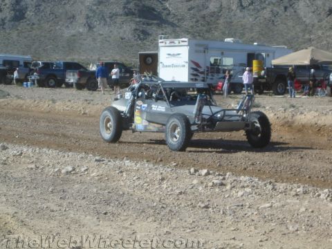 1990 Ford Bronco - dark tube-frame off-road buggy speeds along a banked dirt track with RVs, trucks and spectators lining the background.