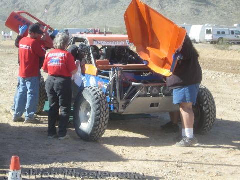 1990 Ford Bronco - pit crew in red shirts works on a tube-frame off-road buggy with an open orange rear cover and BFGoodrich tires in the desert.