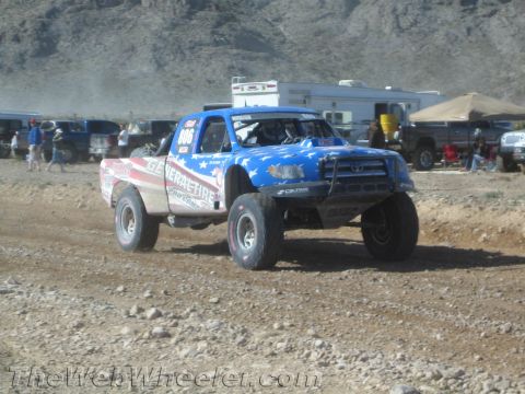 1990 Ford Bronco - off-road race truck numbered 906 with a stars-and-stripes hood catches air on a dusty desert course near RV haulers.
