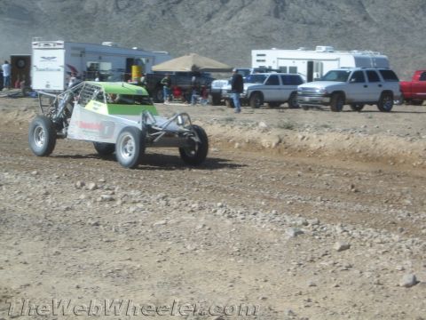 1990 Ford Bronco - green and white single-seat tube-frame desert buggy idles on a dirt track in front of parked toy haulers and SUVs.