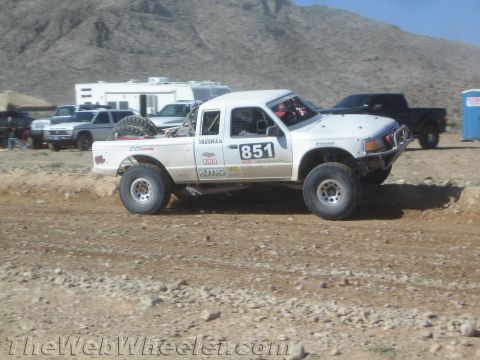 1990 Ford Bronco - white off-road race truck numbered 851 sits on a dirt course in a desert pit area with chase trucks and a portable toilet behind.