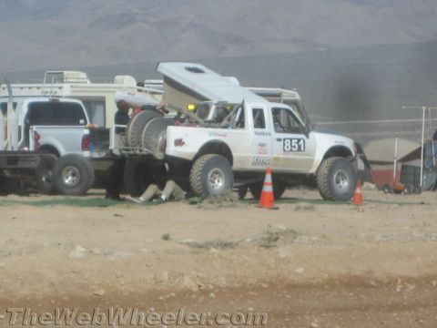 1990 Ford Bronco - Race truck no. 851 sits in a desert pit with its hood propped open while crew members work on it beside chase trucks.