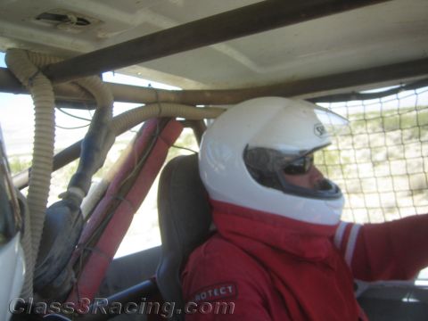1990 Ford Bronco - driver in a white full-face helmet and red firesuit sits belted inside the roll-caged cockpit of an off-road race vehicle.