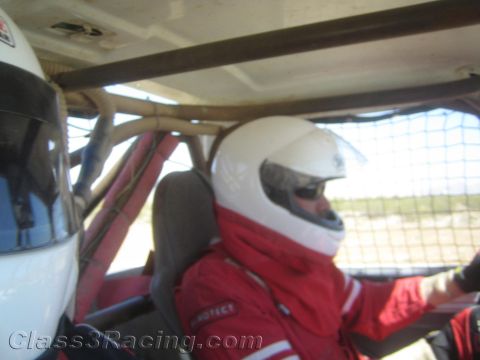 1990 Ford Bronco - Two occupants in white helmets and red firesuits sit side by side in the roll-caged interior of an off-road race vehicle.