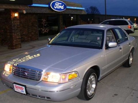1999 Ford Crown Victoria - A silver sedan is parked outside a dealership with a Ford sign, with another vehicle visible in the background.