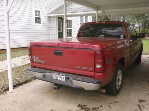 2003 Chevrolet Silverado 1500 - A red Chevrolet Silverado pickup truck is parked in a driveway under a carport, showing its rear end and side.