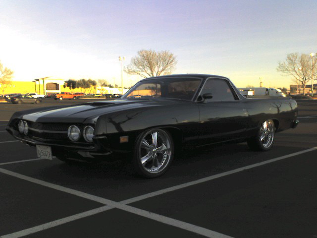 Black Ford Ranchero with hood scoop and polished five-spoke wheels in a parking lot at dusk
