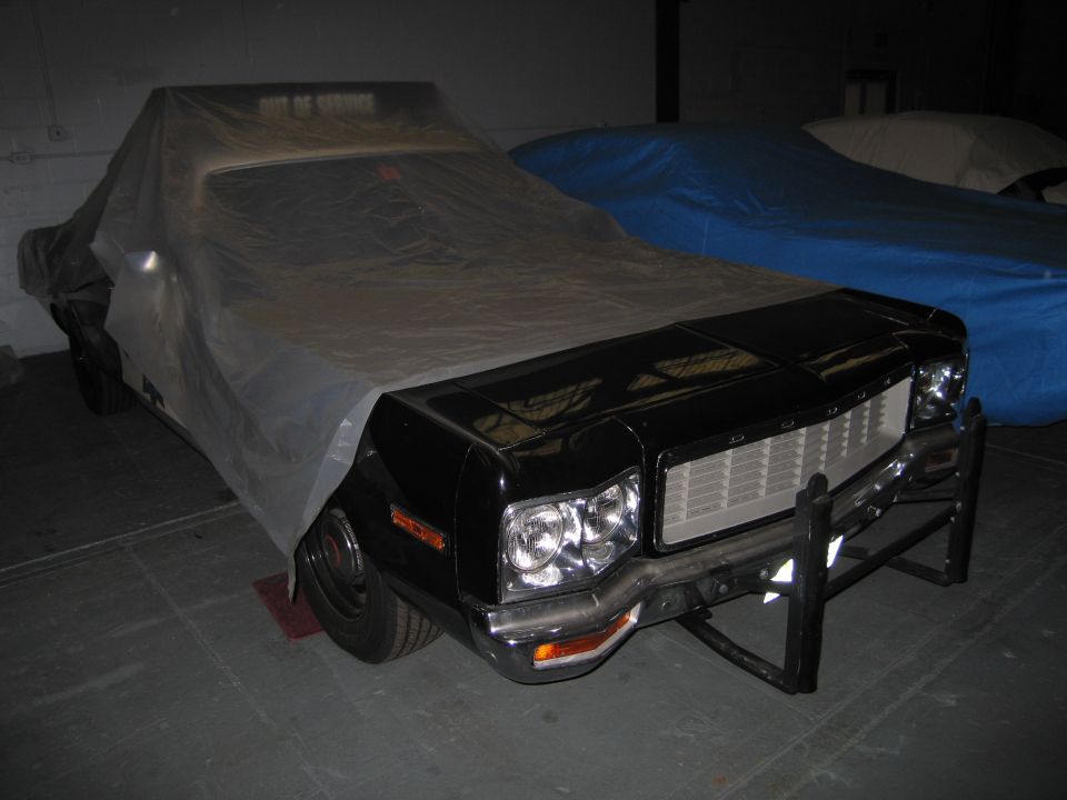 Black car with a push bar, covered in a clear plastic sheet, parked next to a blue car cover