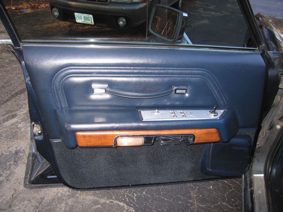 Interior door panel with blue upholstery, wood trim, and power window controls. Another vehicle is visible through the window