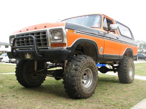 1979 Ford Bronco - Lifted orange-and-black full-size Ford Bronco with a removable hardtop, shot from a low 3/4 front angle on grass. It rides on oversized Sup…