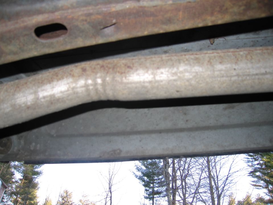 Underside view of a rusty exhaust pipe and frame rail, with trees visible in the background