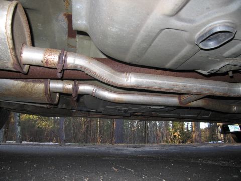 1991 Ford LTD Crown Victoria - The undercarriage of a vehicle, its exhaust system and fuel tank, with a wooded background.