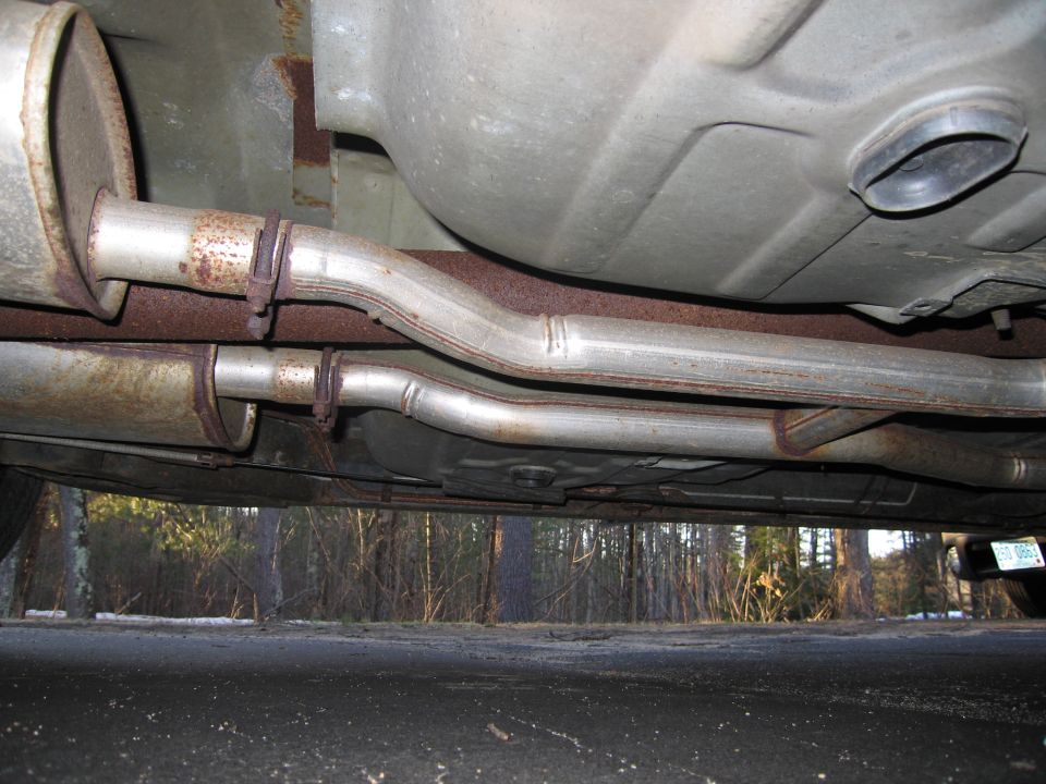 Underside of a car showing dual exhaust pipes and mufflers. A fuel tank is visible above