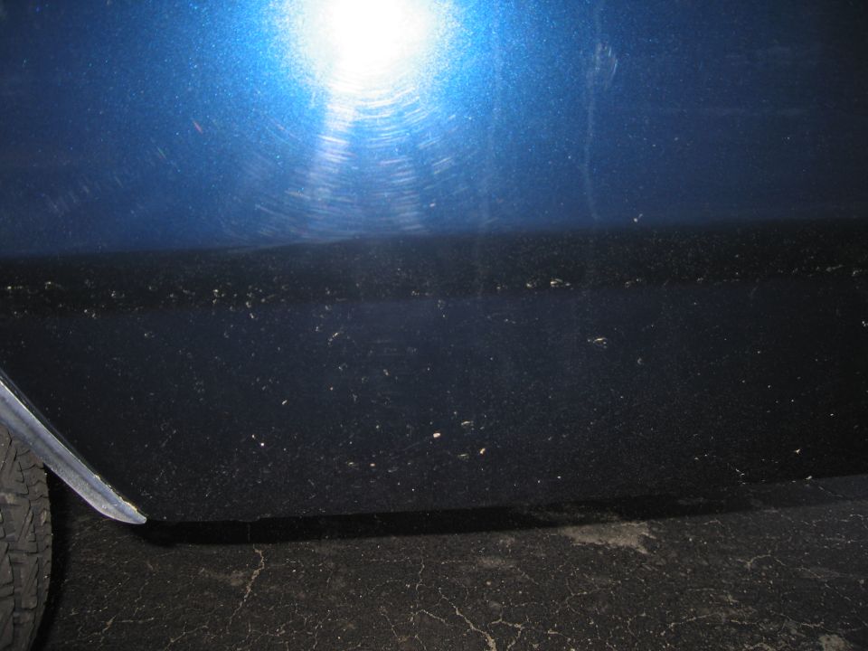 Close-up of a blue car's side panel with some dirt and scratches. A tire is partially visible on the left