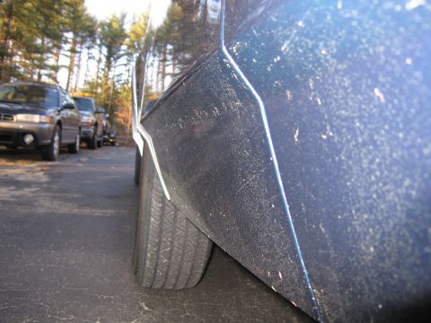 1991 Ford LTD Crown Victoria - A close-up side view of a dusty blue car's rear quarter panel and tire, with other vehicles parked in the background.
