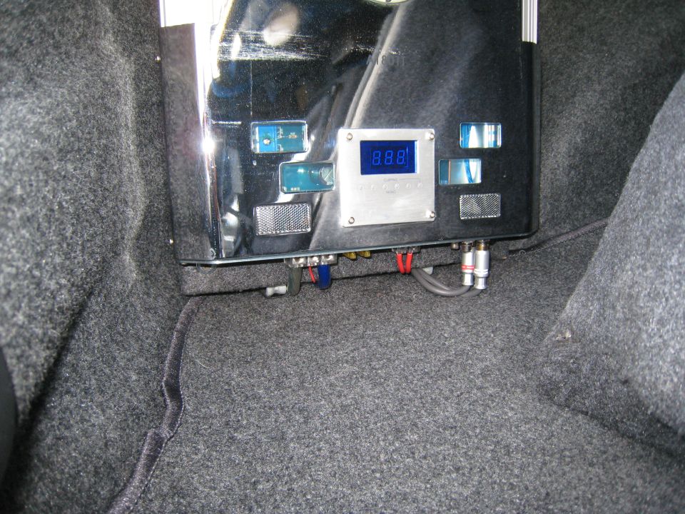 Car audio amplifier with blue digital display and RCA connections, mounted in a carpeted trunk