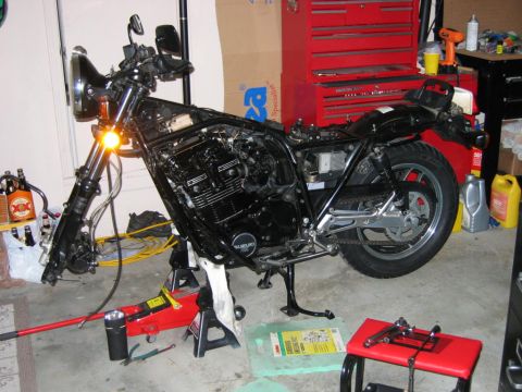 1985 Ford LTD Crown Victoria - A motorcycle is disassembled and supported by jack stands in a garage, with tools and beverages present.