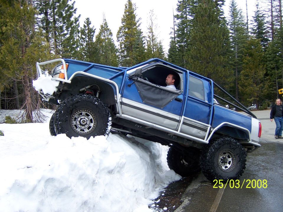 Blue pickup truck with roll cage and large tires climbing a snow bank