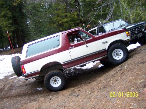 1991 Ford Bronco - Two Ford Broncos navigate a snowy, wooded trail on a dirt incline.