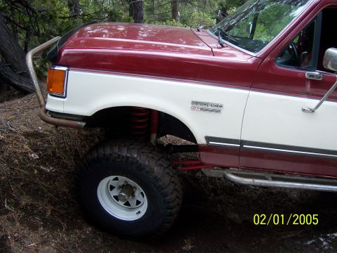 1991 Ford Bronco - A two-tone red and white Ford Bronco is shown off-road with large tires and a custom brush guard.