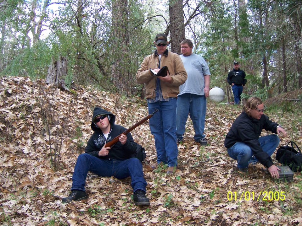 Group of men in a forest with one holding a rifle, timestamped 01/01/2005
