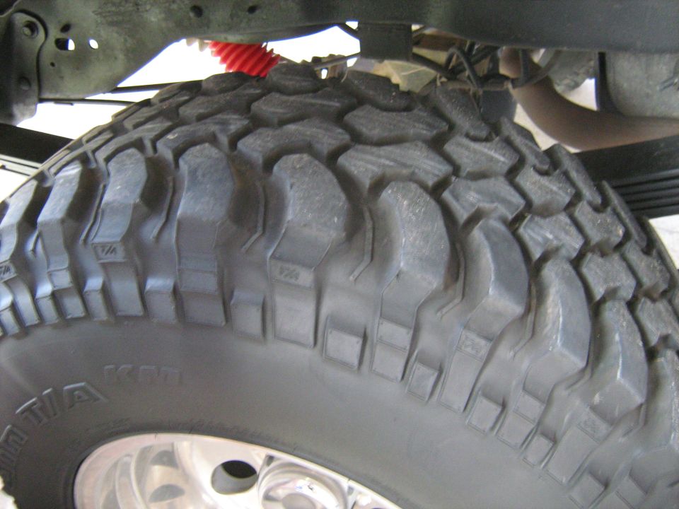 Close-up of a large off-road tire with aggressive tread, showing suspension components and exhaust pipe above
