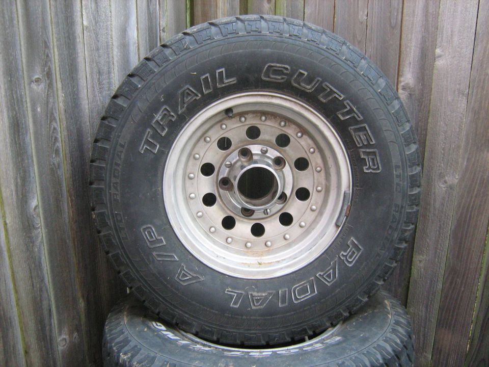Tire with 'Trail Cutter' text and a silver wheel with multiple lug nut holes, leaning against a wooden fence