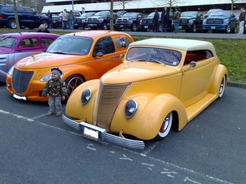 1991 Ford Crown Victoria - A yellow vintage coupe is parked next to an orange PT Cruiser, with a child standing in between.