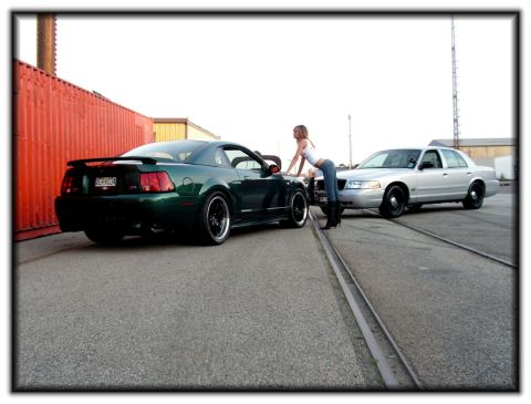 1998 Ford Crown Victoria - A woman leans on a green Mustang next to a silver 1998 Ford Crown Victoria parked on railroad tracks.