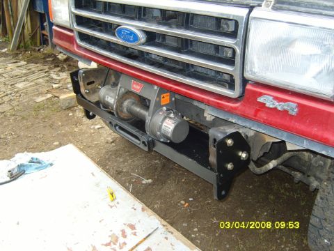 1989 Ford Bronco - A Warn winch mounted on a black tubular winch bumper being test-fitted to the front of a red Ford truck, with the mounting brackets bolted…
