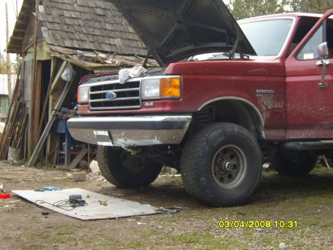 1989 Ford Bronco - A red 1989 Ford Bronco sits with its hood open, parked in front of a rustic wooden structure.