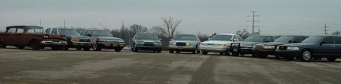 1998 Ford Crown Victoria - A collection of police cruisers and older sedans are parked in a lot under an overcast sky.