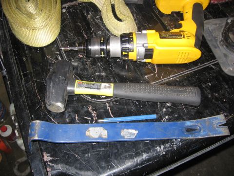 1989 Ford Bronco - A collection of tools including a yellow drill, sledgehammer, crowbar, and a yellow strap are laid out on a dark, textured surface.
