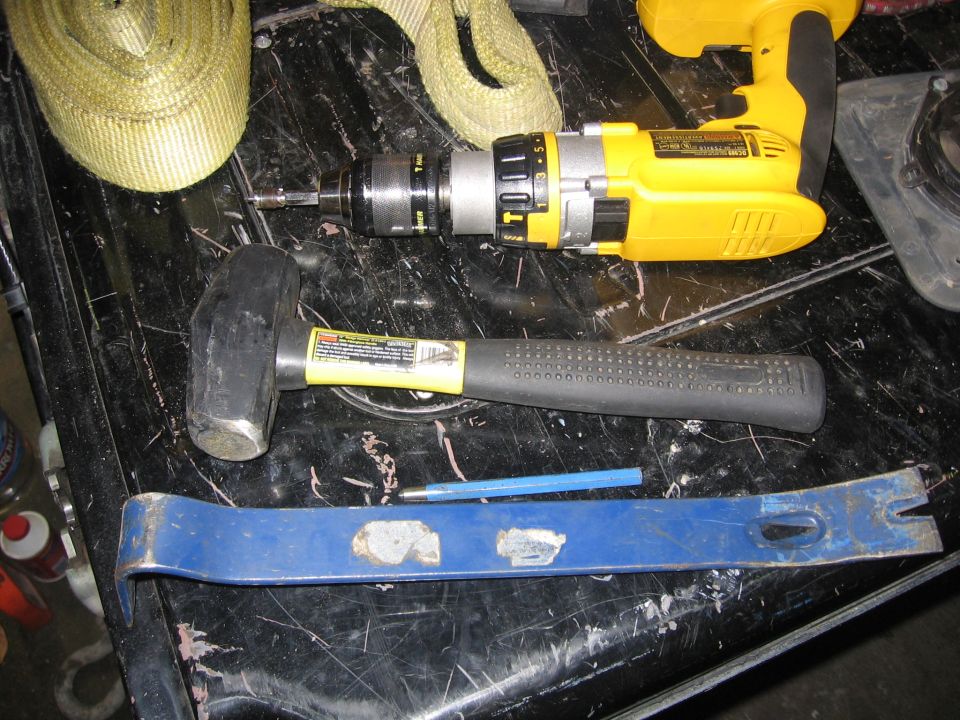 Yellow drill, sledgehammer, crowbar, and strap on a dark surface
