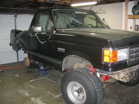 1989 Ford Bronco - A black Ford Bronco is shown in a garage with its front end raised on hydraulic jacks, revealing a cracked windshield.