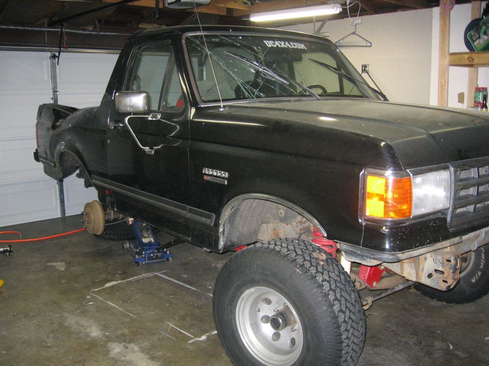 Black Ford Bronco in garage, front end lifted on jacks, cracked windshield with sticker that says DC4X4.COM