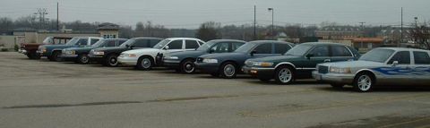 1998 Ford Crown Victoria - A row of sedans, some appearing to be police or taxi vehicles, are parked in an outdoor lot.