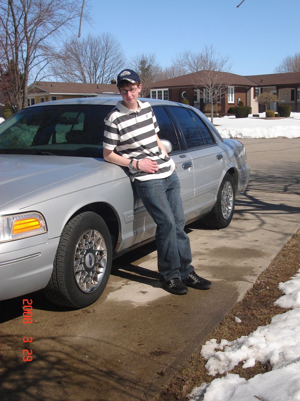 Man leaning on the front fender of a silver Ford Crown Victoria sedan in a snowy driveway