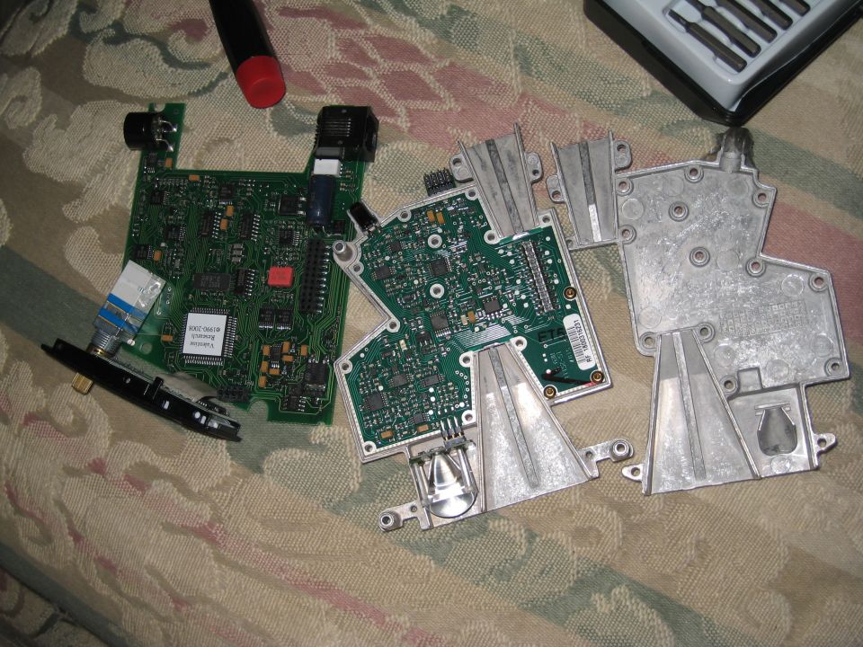 Disassembled electronic circuit boards and metal casing parts on a patterned fabric surface. A screwdriver is visible at the top left