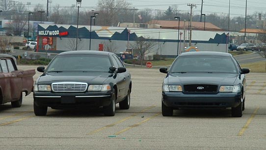 Two black Ford Crown Victorias parked in a lot, one with a chrome grille, the other with a black grille