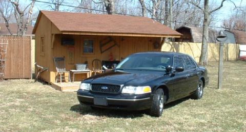 1998 Ford Crown Victoria - A black sedan is parked in a grassy yard next to a small wooden shed.