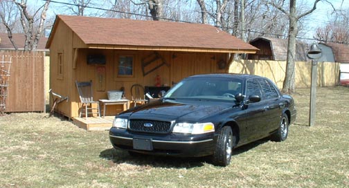 Black sedan parked in front of a wooden shed. Watermark says SUPERMOTORS