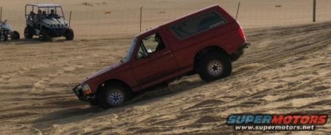 1984 Ford Bronco - A red SUV is parked on a dirt surface, with another vehicle blurred in the background.