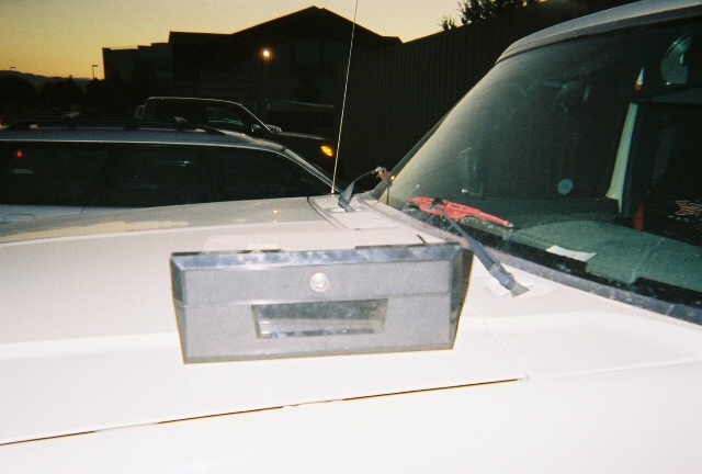 White vehicle hood with gray storage box and antenna; another vehicle is parked behind