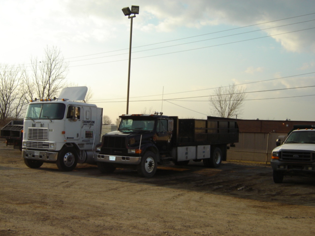 Two trucks parked on dirt lot, one white cab-over, one black flatbed