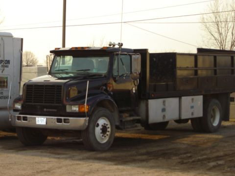 1992 Ford Bronco - A dark blue flatbed truck is parked next to a white trailer. The truck has a black bed and chrome accents.