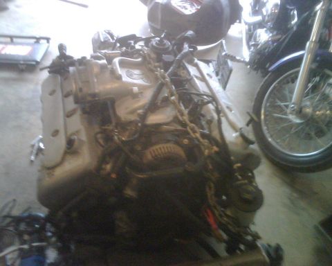 1999 Ford Mustang - Engine assembly with visible wiring and chains, situated on a concrete floor.