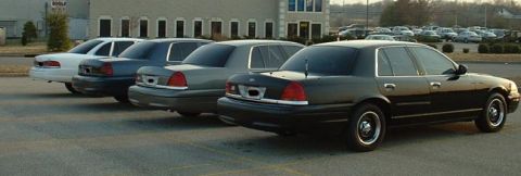 1998 Ford Crown Victoria - A line of four sedans, including a black 1998 Ford Crown Victoria, are parked in an outdoor lot.